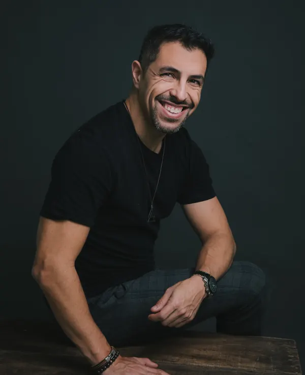 Fernando Souza sorrindo, sentado à mesa de madeira no estúdio.